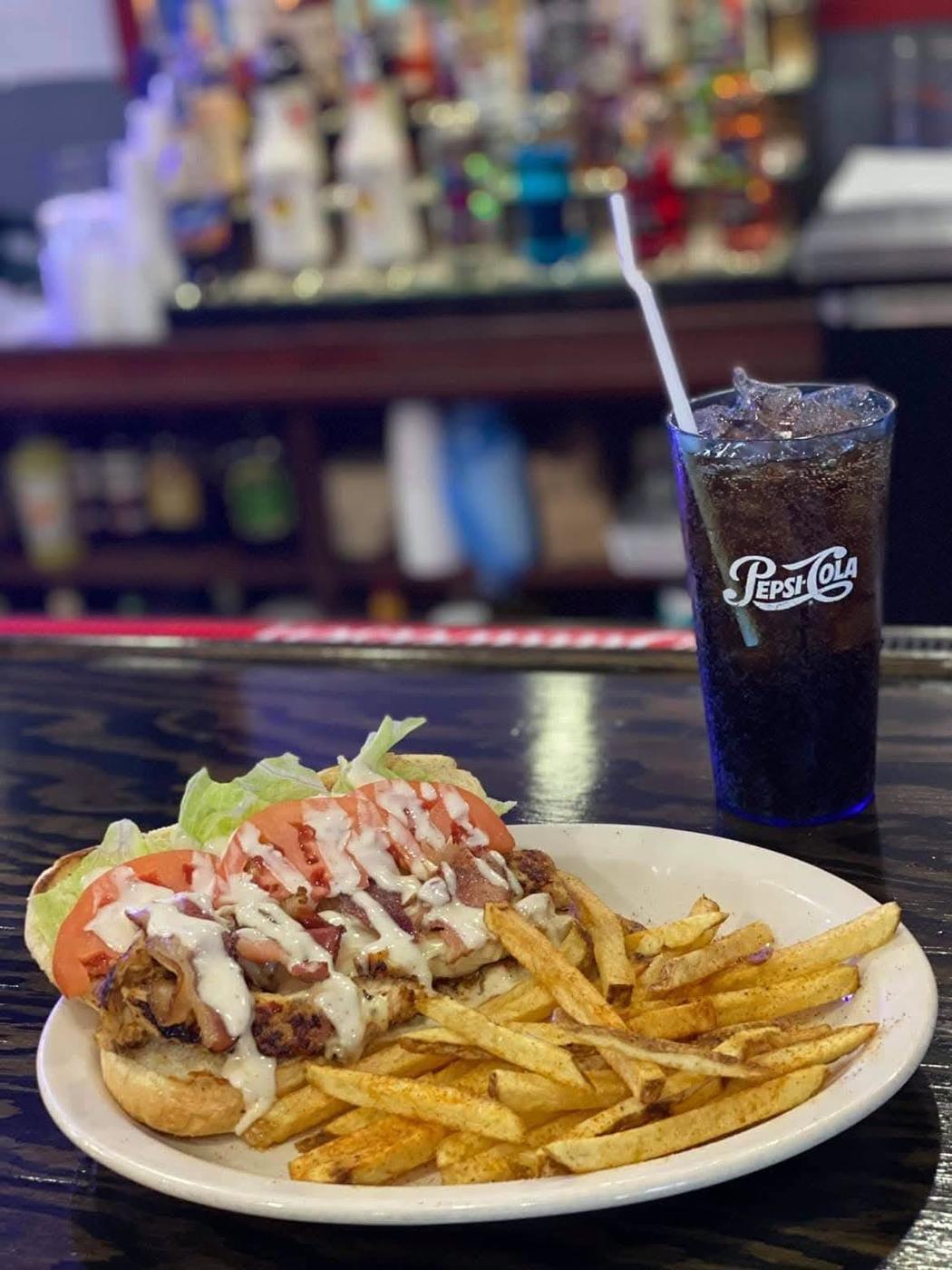 A chicken sandwich with bacon, lettuce and tomato beside seasoned fries, on the bar top with the back bar bottles out of focus behind