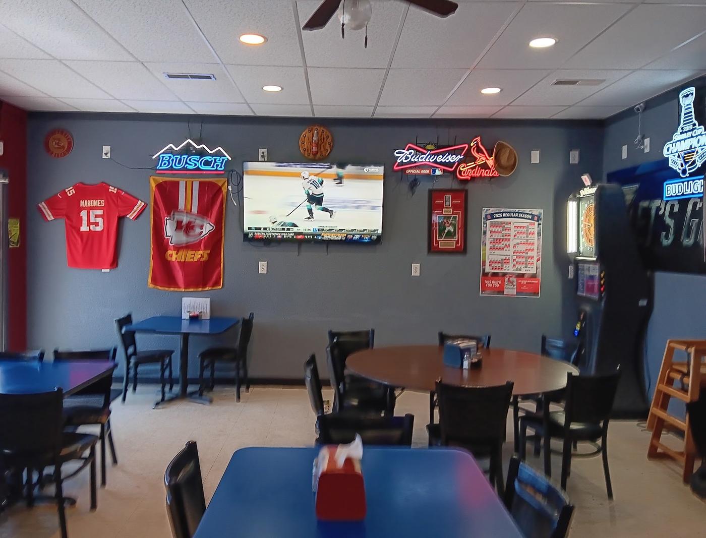 The dining room at West End Bar and Grill: gray and red walls with a framed football jersey and a team flag, a wall mounted TV, a lit dart machine and blue and wood tables with black chairs